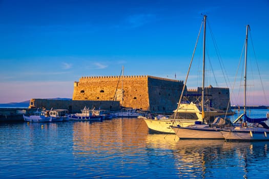Landscape of Venetian castle or Koules fort in Heraklion, capital of Crete island, Greece and moored fishing boats at sunset. Clear blue sky, deep blue waters of port, yachts, sailboats. Travel destination of Mediterranean sea, summer vacations.
