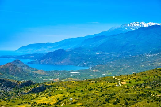 Summer day landscape of Crete island, Greece. High mountains, snow-covered peaks, green hills, roads and olive tree plantations, high grass, lush foliage and vegetation. Mediterranean sea, blue sky and clouds. Idyllic panorama on sunny day.