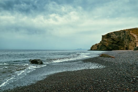 Dramatic landscape of stormy sea or ocean waves rolling on rocky volcanic beach on cloudy day. Overcast sky, heavy grey clouds, black lava pebbles and sand, weathered cliff. Santorini island, Greece.