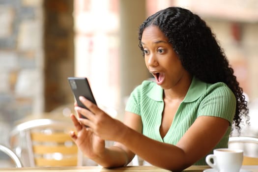 Amazed black woman checking smart phone sitting in a bar terrace