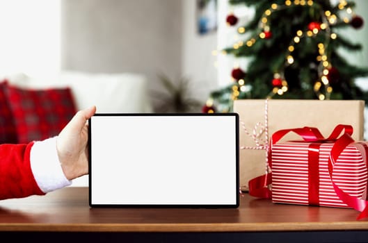 Christmas, New Year background with copy space for design. Santa hand holding empty blank white tablet screen. Mockup for product placement montage. Online shopping from home. Fir tree on background