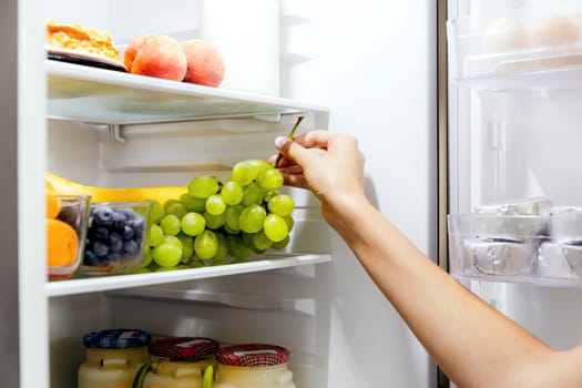 Woman hand taking, grabbing or picks up green bunch of grapes out of open refrigerator shelf or fridge drawer full of fruits, blueberries, bottles with yogurt. Healthy food diet, lifestyle concept