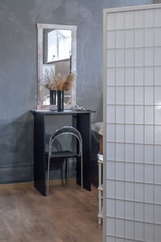 Modern room furnished with white folding screen, rectangular mirror hanging over black table where vase of beige pampas grass standing. Comfortable place with laminate floor and gray walls. Vertical.