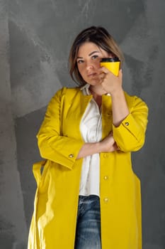 Glorious young woman wearing bright yellow coat over white shirt and blue jeans standing against gray wall with coffee cup in hand. Brunette holding hot drink in paper bowl close to face. Vertical.