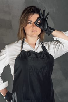 Confident young woman in sable apron over white shirt standing against gray background and showing ok sign close to face. Lady with brown short hair in latex gloves looking through fingers. Vertical.