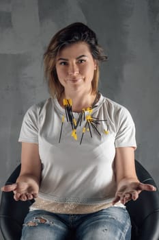 Graceful young brunette sitting on black chair on gray background and throwing up yellow eyelash extension brushes. Lashmaker in white t-shirt smiling, small equipment flying in air. Vertical.
