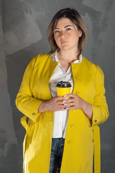 Delightful young woman in bright yellow coat standing against gray background indoors with coffee cup in hands. Relaxed lady with brown short hair holding hot drink and looking ahead. Vertical.