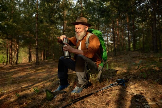 Senior man hiker drinking warm tea from thermos cup during rest in forest. Hiking outside on retirement