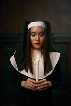 Portrait of beautiful nun holding burning candle in hands while praying. Vow of obedience, innocence and atonement for sins concept