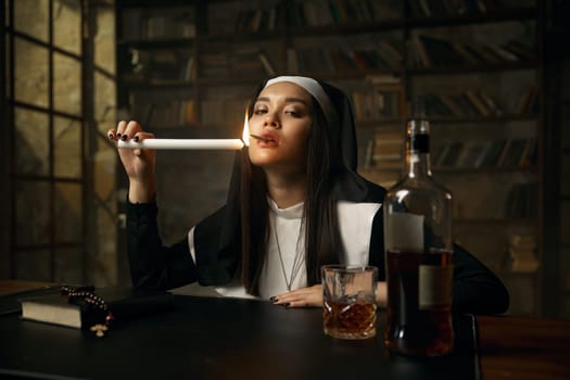 Immoral christian nun lighting cigarette from burning candle sitting at table with opened bottle of whiskey. Portrait of vicious woman dressed in monastic costume