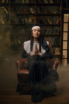 Young beautiful woman in nun dress smoking cigarette sitting on armchair with folded hands in pray position. Attractive holy sinner portrait