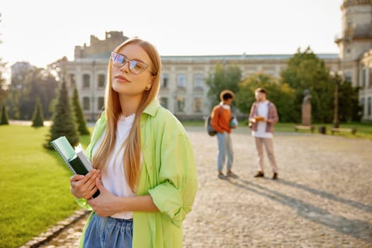 Outdoors portrait of smart female student wearing eyeglasses and casual clothes holding studying accessories standing over university campus yard with classmates on background