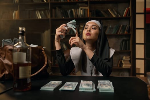 Pretty confident woman in nun dress sitting at table with money dollar cash stacks, whiskey and smoking cigarette. Religion and business, earnings in intimate service field concept