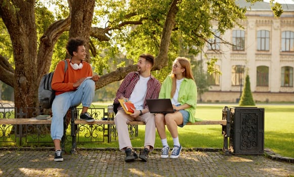 Confident smiling university students chatting together while taking rest at outdoor campus park. Nice conversation of international millennial people group
