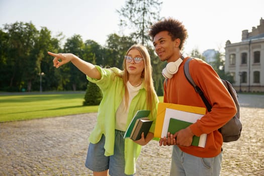 Male and female students gossiping together standing at university campus backyard. Teenage boy newcomer asking millennial girl for help to find education class or library