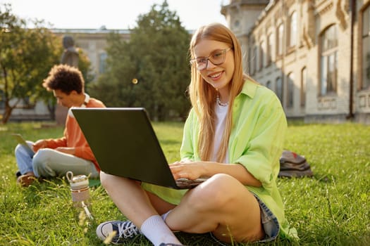Portrait of smart millennial female student happily smiling using laptop computer while studying outdoors with classmates at university campus park. Back to education concept