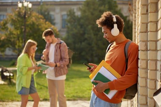 Teenager students at university campus after day of class. Young guy in headphones using mobile phone standing with textbooks in foreground and classmates friendly talking on blurred backdrop