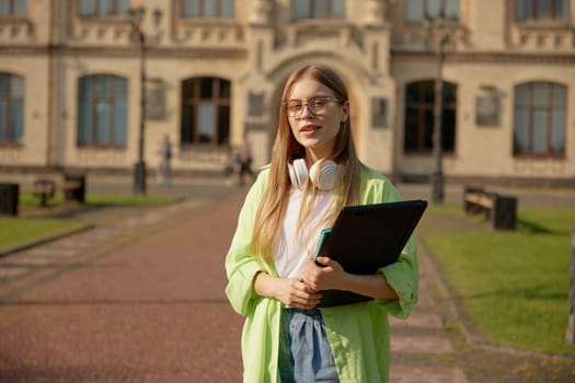 Portrait of teenage female student over university building on background selective focus. Confident casual young girl carrying laptop computer