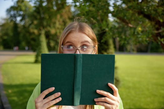 Portrait of cute funny female student hiding behind book looking at camera with amazed, shocked facial expression