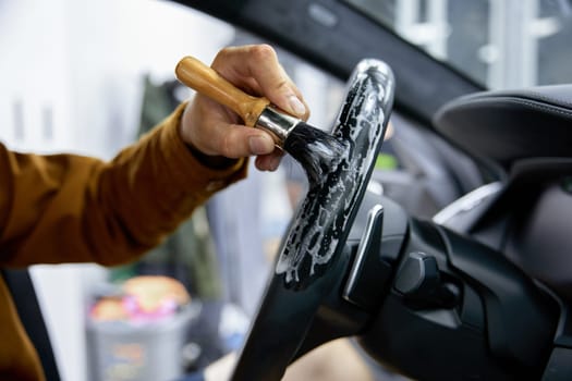 Car detailing and dry cleansing service. Closeup worker hands holding brush. Male technician cleaning leather steering wheel from dirt and dust with special foam