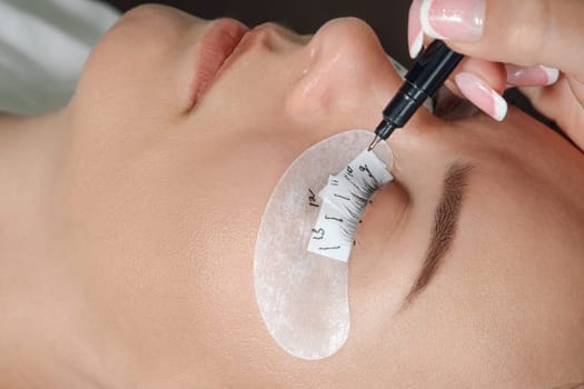 Close up of face of young woman with white thin patches on lower eyelid and closed eyes calmly lying while lashmaker marking places to gluing artificial eyelashes with black pen.