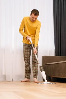 Mature human wearing comfortable clothes washing floor in living room near couch. Man in yellow longsleeve, plaid pants and barefoot standing close to curtains and wiping ground. Vertical.