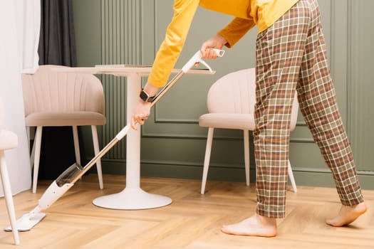 Unrecognizable human in plaid pants and yellow longsleeve, watch on hand washing floor using mop near round table at home. Housekeeper in comfortable clothes and barefoot cleaning room.