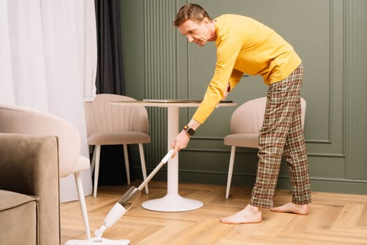 Thin mature human wearing comfortable clothes washing floor in kitchen near round table using mop. Housekeeper in yellow longsleeve and plaid pants wiping ground from dust.