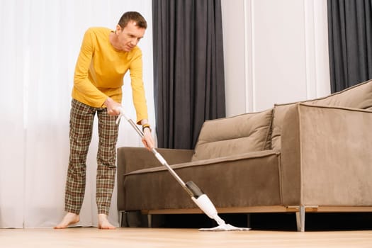 Middle aged man wearing home clothes wiping floor in living room near beige and grey couch. Male housekeeper cleaning room, doing housework using mop in yellow longsleeve and plaid pants.