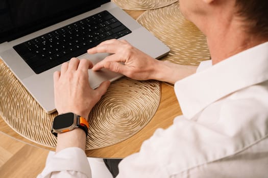 Close up of hands of adult man in white shirt working at laptop at table. Hard-working business man with sporty orange watch on wrist operating on computer indoor.