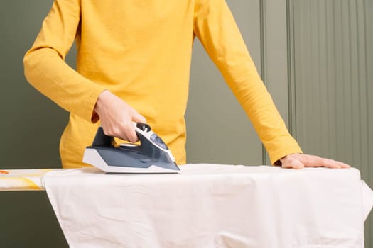 Unrecognizable human in yellow longsleeve standing and ironing white shirt on pressboard using electric flatiron. Thin man doing housework and take care of clothes.