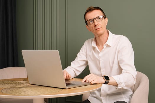 Portrait of middle aged , thin business man sitting at round table on which lying open laptop. Serious looking man in glasses and white shirt distracted from work at comfortable home.