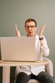 Stunned middle aged man furiously raised hands while working on laptop from home office. Workman in glasses dissatisfied with work, bemused by result, look at computer monitor while sitting at table.