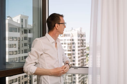 Mature thin human in glasses wearing white shirt standing near window with curtains in multi-storey building. Business man with dark hair put hands together and peering into distance from high floor.