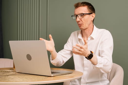 Puzzled mature human sitting at table and looking at laptop screen with misunderstood eyes. Dazed business man in glasses and white shirt working from home raised hands in shock.
