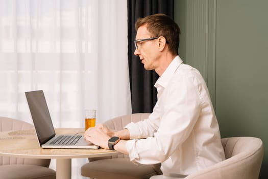 Thin workman in glasses and white shirt working hard at computer sitting at round desk. Serious middle aged business man staring at monitor, busy with case, glass with drink on table.