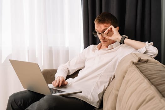 Adult man looking indignantly at laptop screen for which working. Business male operating at home sitting on soft couch and staring in surprise at computer and adjusting glasses with hand.