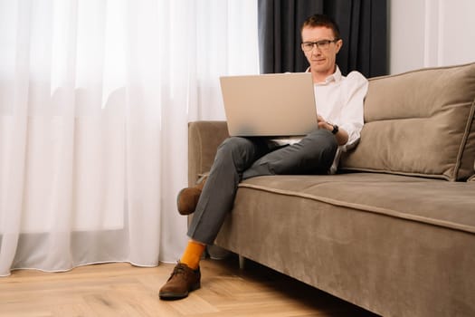 Mature human operating while sitting on brown-grey couch and looking at laptop screen with furrowed brows and disgruntled eyes. Business man dissatisfied with work, working from home office.