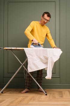 Mature thin man in glasses ironing snow-white shirt on pressboard using flatiron. Soigne tall human in plaid pants, yellow longsleeve and bare feet working around house. Vertical.
