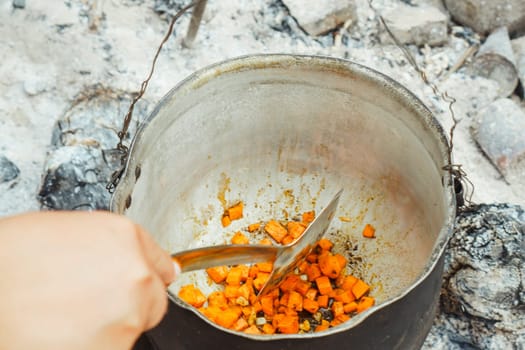 Hand of unrecognizable adult man holding skimmer and stirring cut carrot with spices and seasonings, preparing for cooking meal in metal cauldron on campfire for summer camping day.