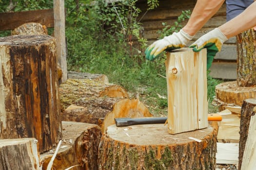 Unrecognizable adult man in gloves standing near countryside house and separating wood on halves with hands after cutting firewood with ax. Tool lying on stump while male preparing tree for campfire.