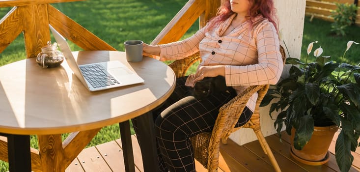 Woman working with laptop outdoor in summer terrace with black kitten. Girl and cat watching video on computer in the evening. Working from home, business, pet and lifestyle
