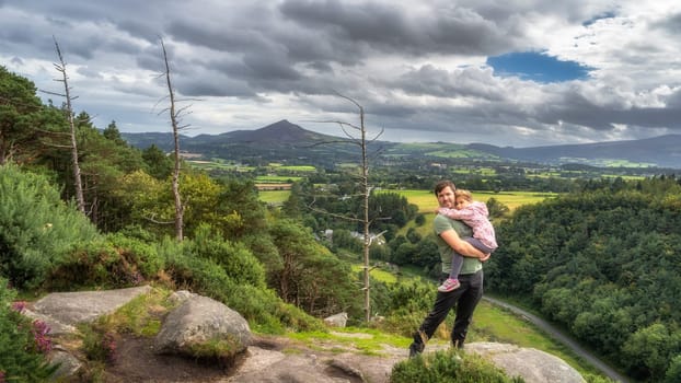 Father holding daughter in arms and standing on edge of cliff, with a scenic view on a valley and pine forest. Family hike, Wicklow Mountains, Ireland