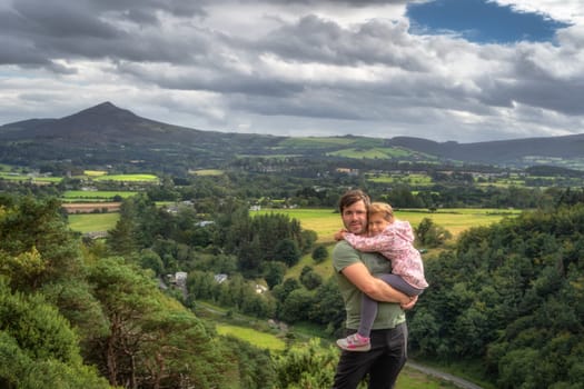 Father holding his daughter in arms with a scenic view on a valley, forest and mountain range. Family hike in Lead Mines, Wicklow Mountains, Ireland