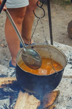 Unrecognizable woman standing near metal cauldron, hanging on hook over firewood, and going to stir savoury soup using skimmer. Lady preparing food on campfire, checking readiness of meal. Vertical.