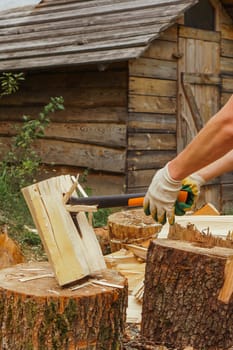 Unrecognizable man in gloves standing near village house and chopping wood, left on stump, using ax. Adult male separating large firewood on halves for campfire in rural area. Vertical.