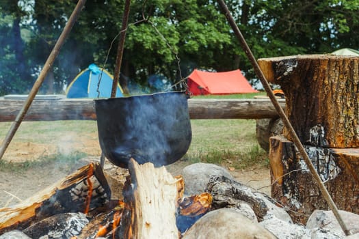 Steam from firewood raising and filling air with delicate and spicy aroma of mouthwatering meal cooking on bonfire in metal cauldron, hanging on tripod in village, preparing for cuisine at camping.