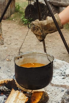 Hand of unrecognizable man wearing glove hanging metal cauldron on hook, leaving vessel with soup on tripod over firewood. Male preparing for cooking meal on campfire, relaxing in village. Vertical.