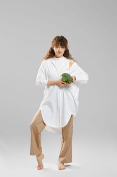 Barefoot slender woman in wide trousers and shirt demonstrates broccoli