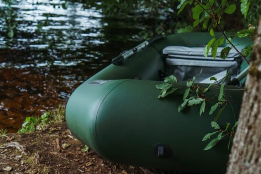 Modern green inflatable boat parked on shore near lake in evening, standing close to tree, slightly covered by branches and leaves. Transportation for floating and relaxing on water in rural site.